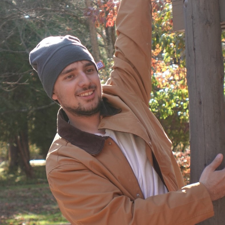 Man in a tan coat and gray beanie standing outdoors with trees in the background