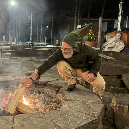 Man tending to a fire pit in a rustic outdoor setting at night wearing a howley polartec hat