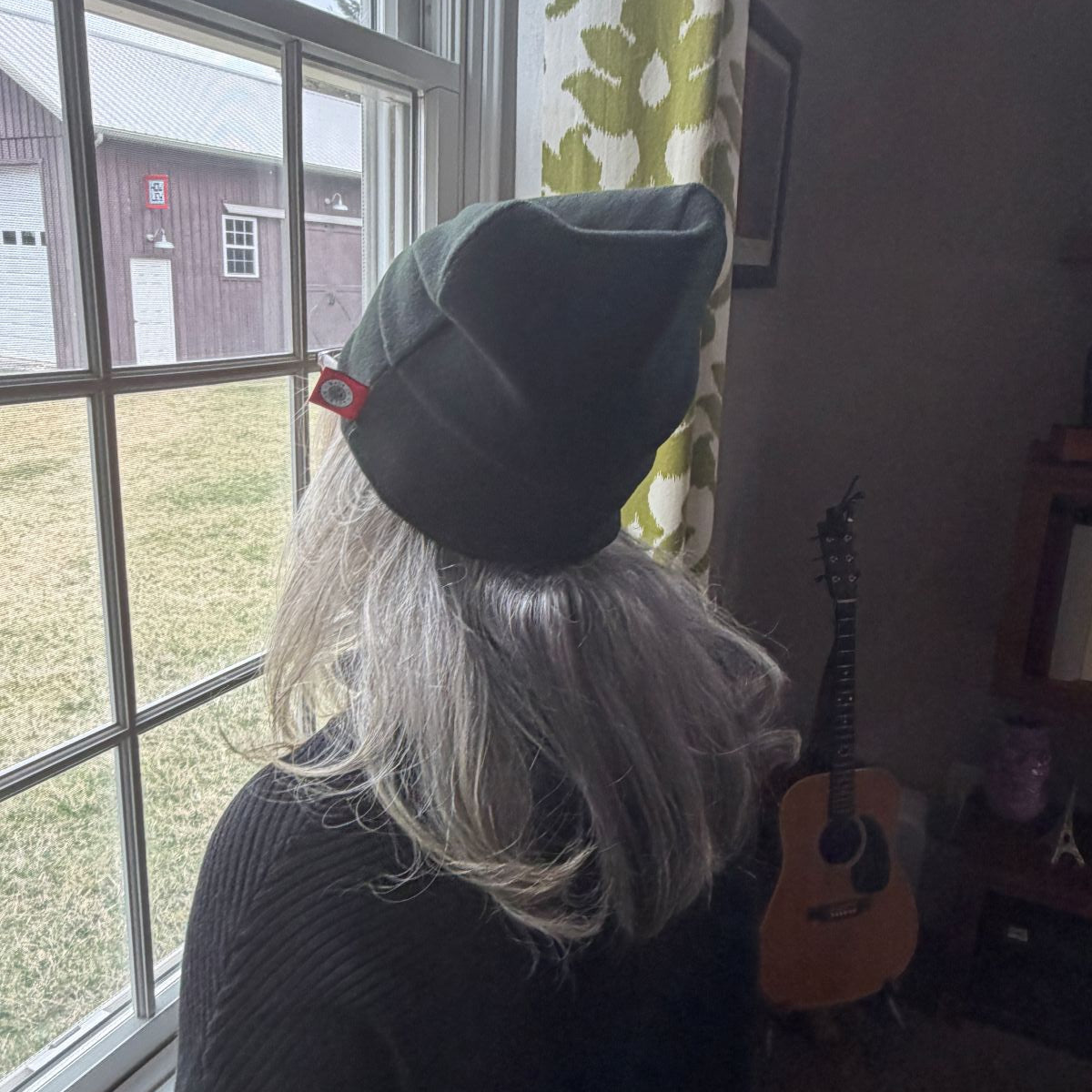 Person wearing a black merino wool beanie with a red tag, sitting by a window with a guitar in the background.